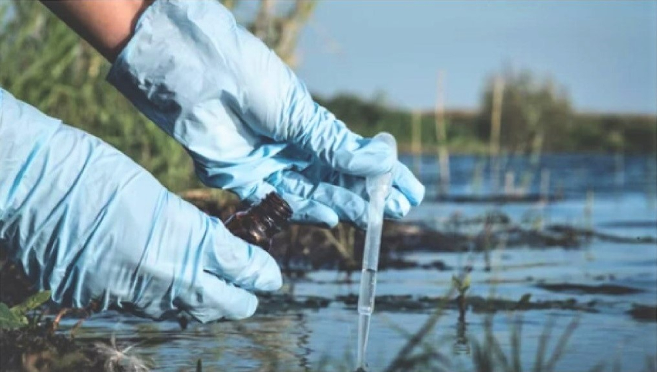 A set of hands wearing rubber gloves is shown with wetlands water and vegetation in the background. One hand is holding an eye-dropper containing drops of water and the other hand is holding a vial that is being positioned to receive water from the eye-dropper.
