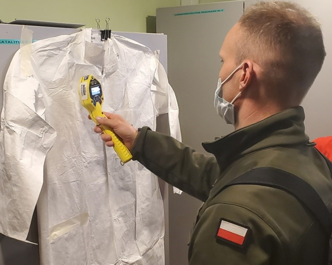 A man in a military uniform with a Polish flag uses a hand-held Geiger counter to examine a hanging dress shirt.