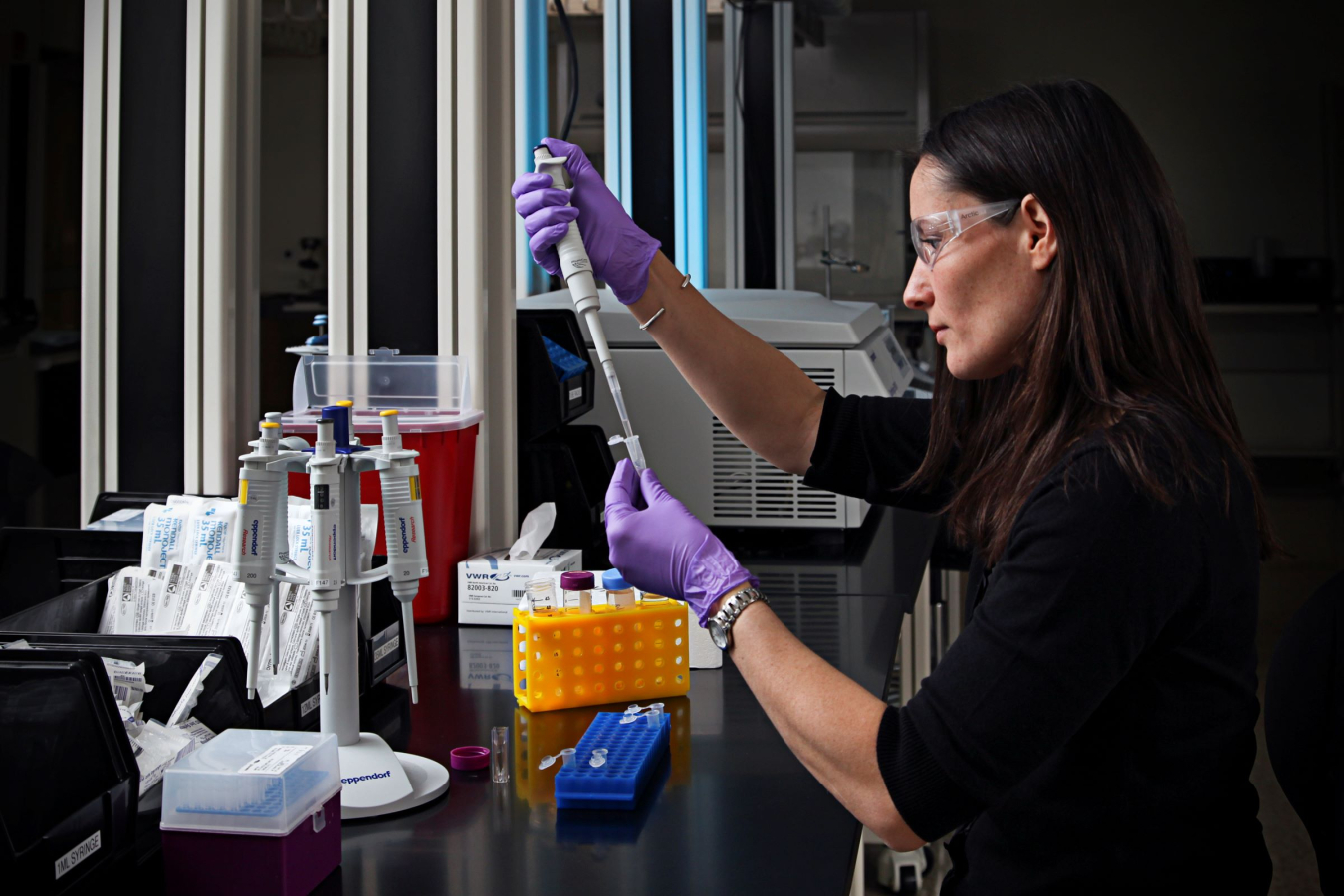 A scientists wearing goggles and purple gloves uses a pipette to transfer liquid into a tube they are holding