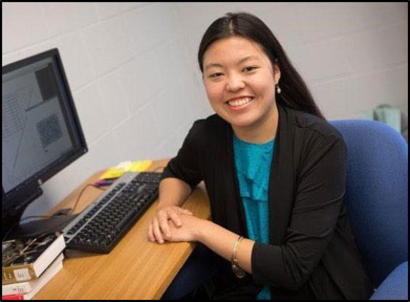 Young woman sits a desk with a computer sitting atop it. Her body is turned toward the camera with her hands draped over one another on the desk. She is wearing a blazer with a teal shirt underneath and smiling at the camera.