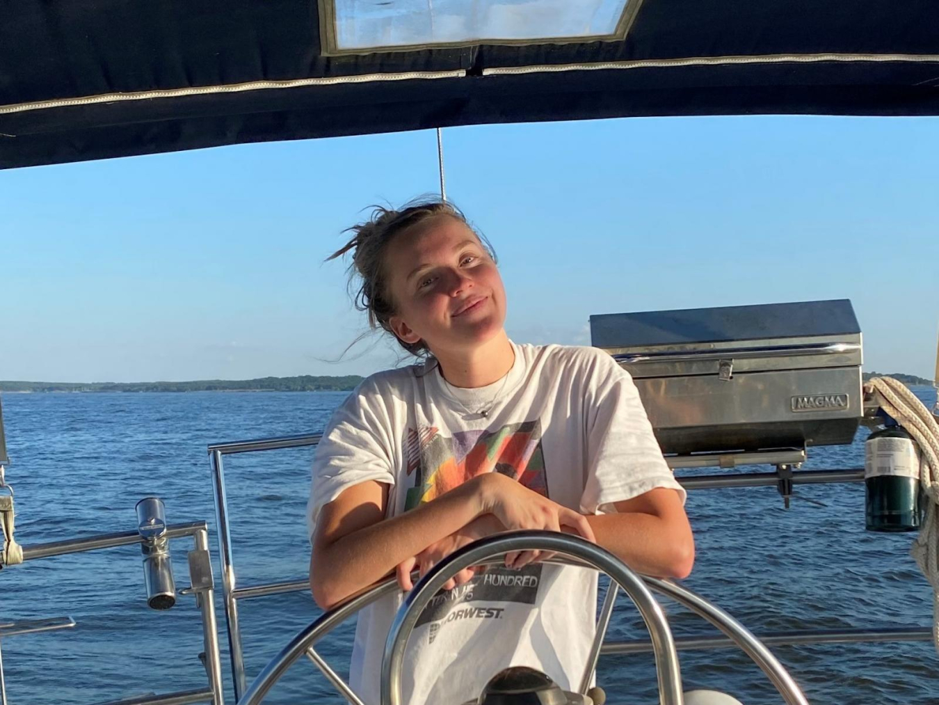 Young woman stands at the wheel of a boat with her arms crossed, surrounded by blue ocean water. She is wearing a white t-shirt, her windblown hair is in a bun, and she is grinning at the camera with her head tilted slightly.