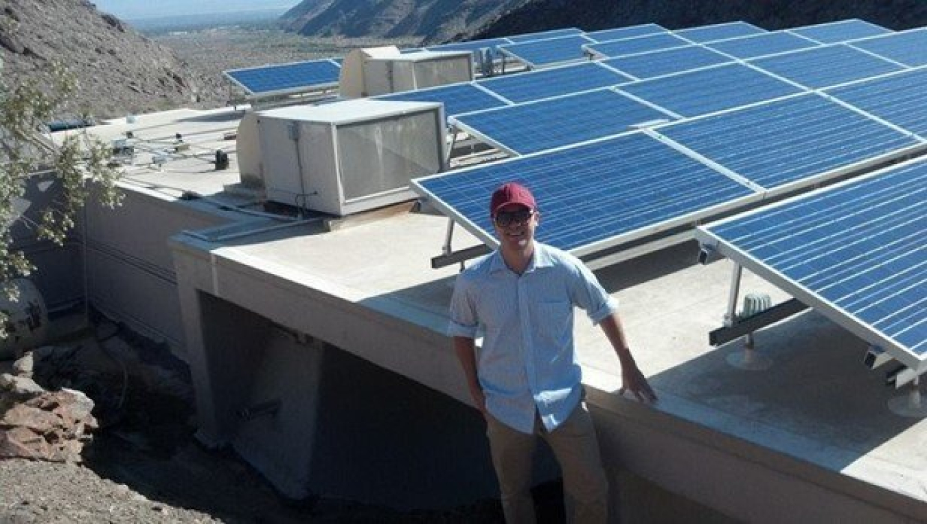 Young man dressed in a button up shirt, khakis, and a red baseball cap leans against a foundation supporting multiple solar panels.