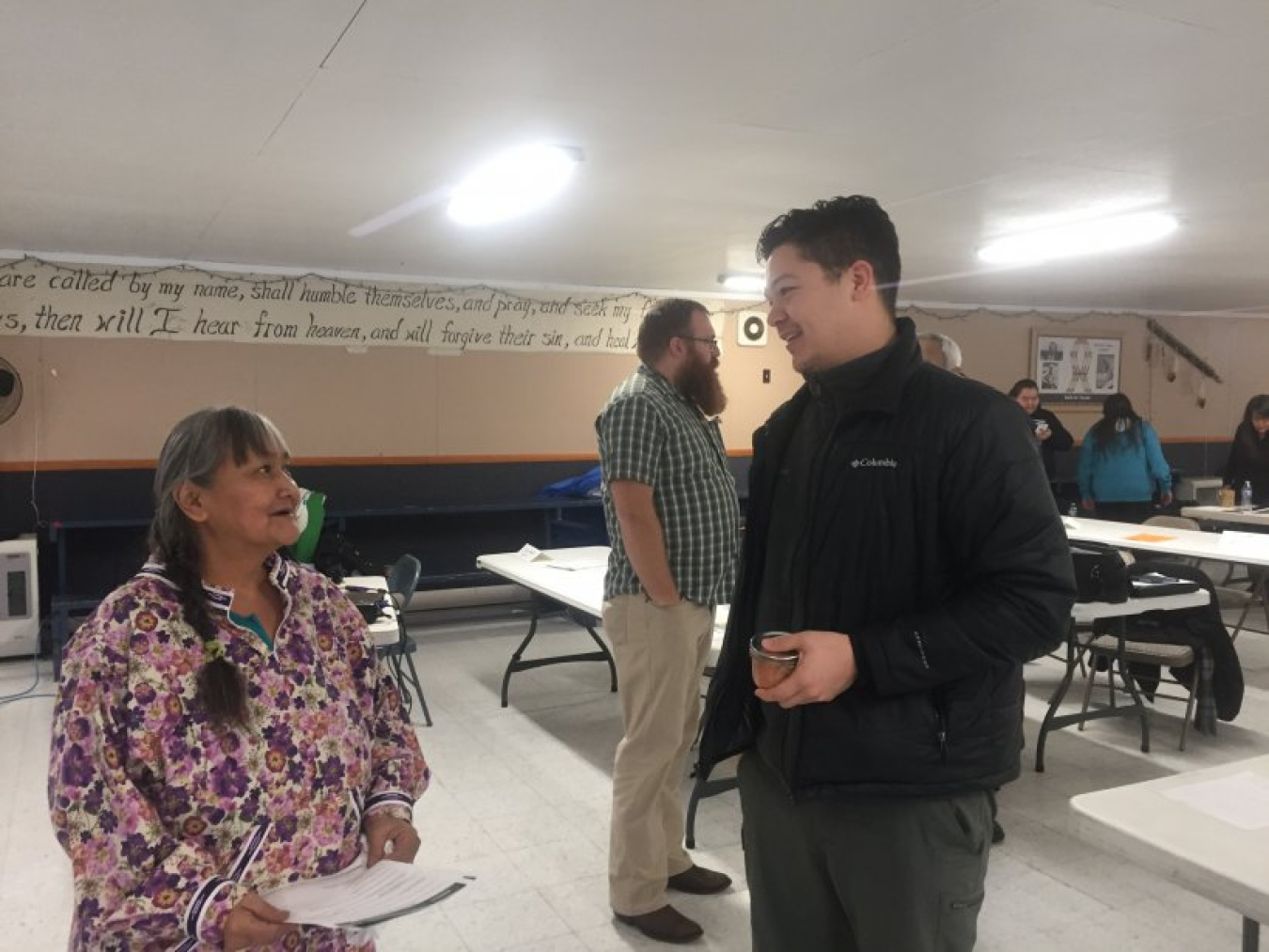 Young man in black jacket talks while smiling to a woman wearing a bright floral shirt and braided hair. They are in a room with multiple white tables and there are other people in the background.