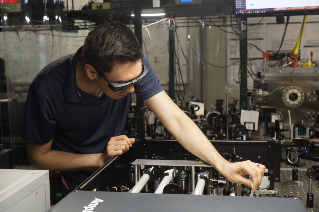 Young man works in a laser laboratory. The room is grey with wires hanging throughout. The man is wearing safety glasses and has one hand resting on a machine's casing and one arm outstretched into the machine working on small objects with his fingers.