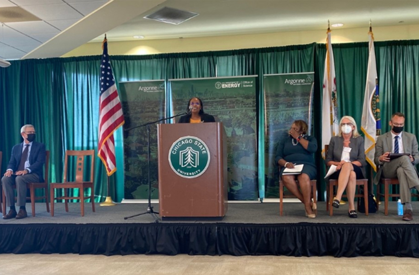 Photo of Dir. Berhe standing at a podium next to an American flag on a stage with people in suits and masks sitting in chairs on both sides of her