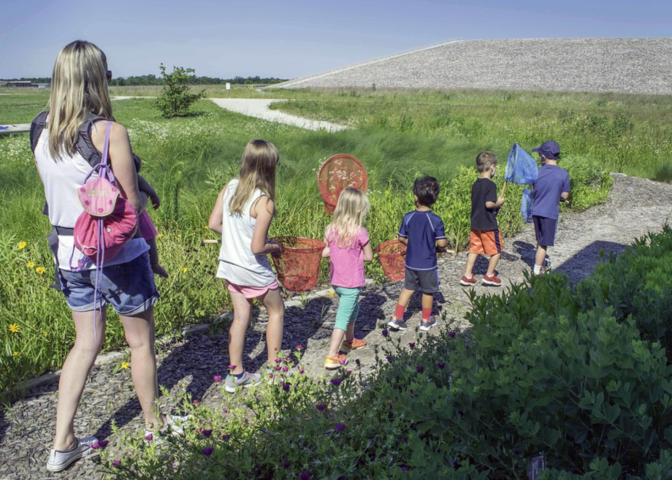 Five children holding butterfly nets walk in a straight line on a path surrounded by greenery. A woman holding a baby follows behind them.
