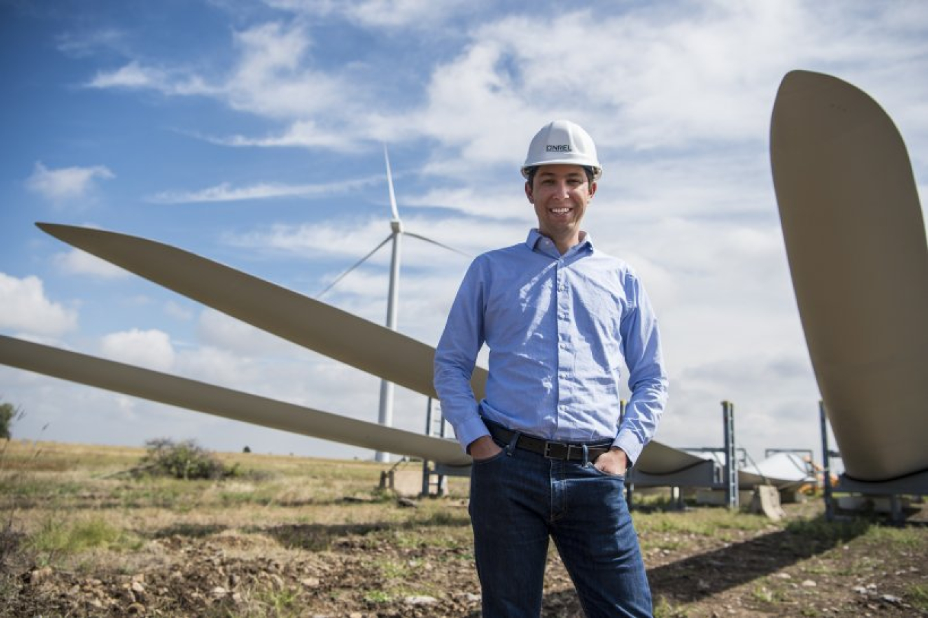 Young man wearing light blue button up shirt, jeans, and a hard hat smiles at the camera with his hands in his pockets. He stands in front of wind turbine blades that are lying on the ground in a grassland setting.