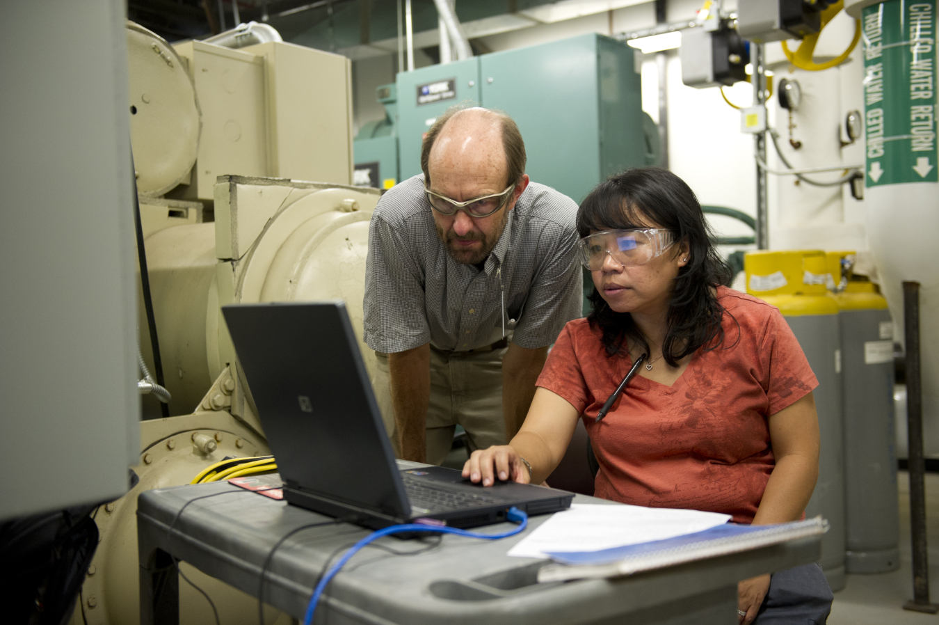 Two people wearing safety glasses looking at a laptop in a utility room.