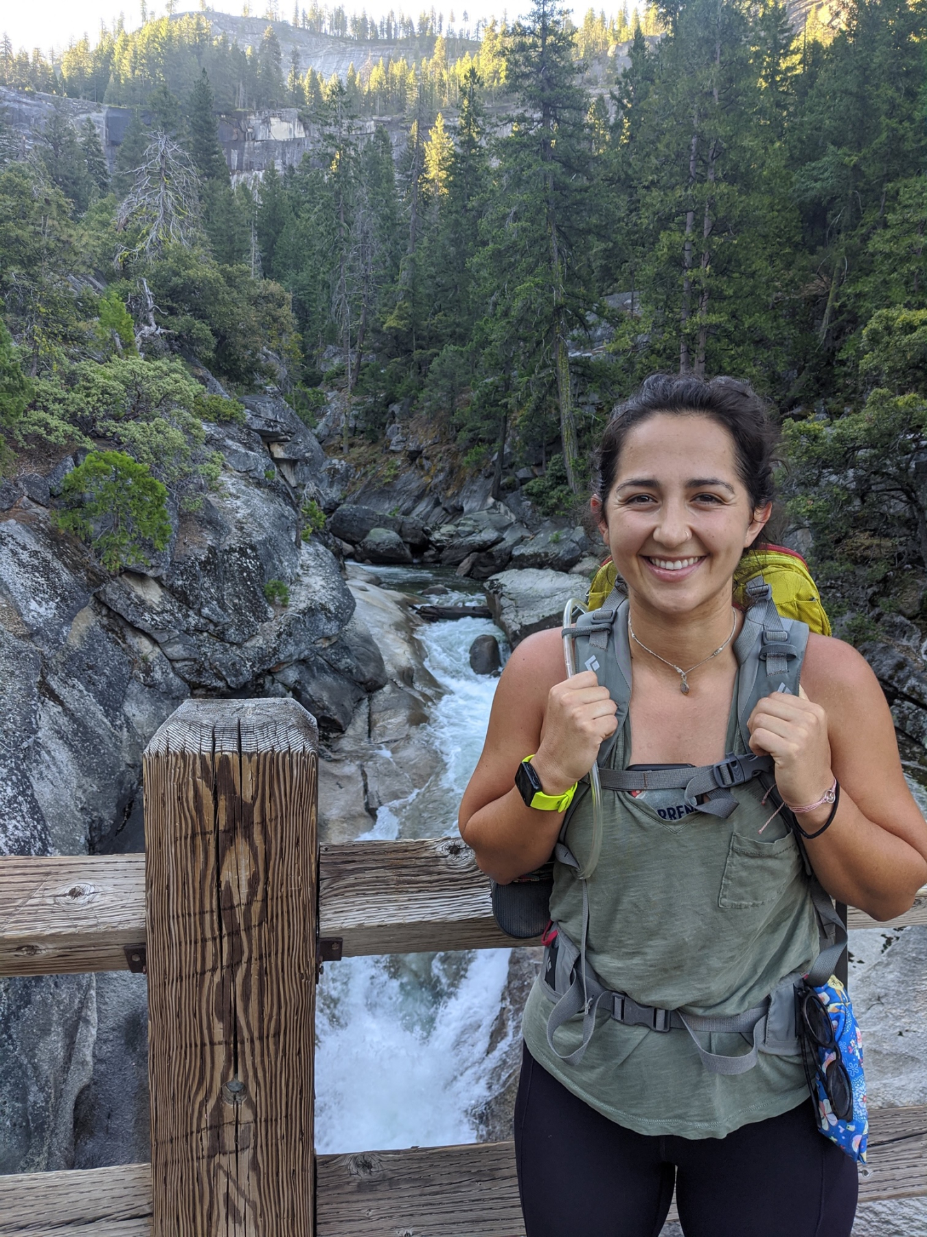 Claire Gonzales, wearing a backpack, stands on a wooden bridge over a river through the woods.