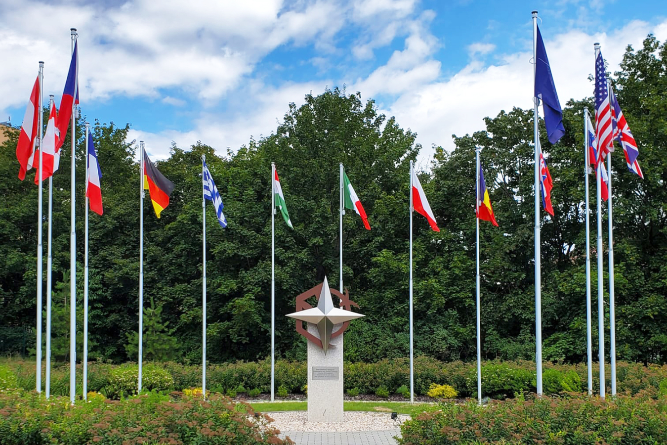 A photo of a park or outdoor space with several national flags in a semicircle surrounding a symbol of NATO sitting on a platform.