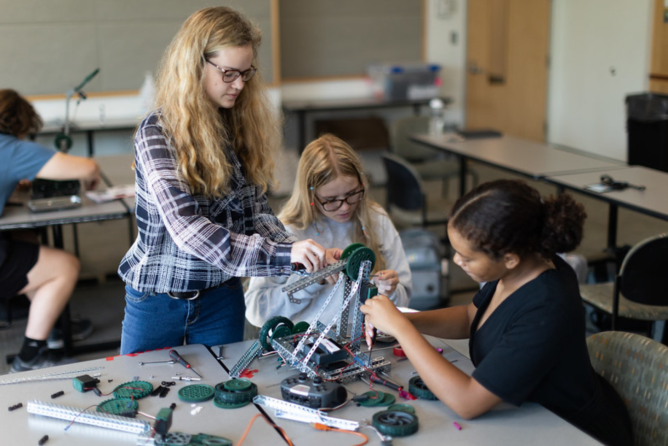 Three young women work together in a classroom setting to build a robotic machine.