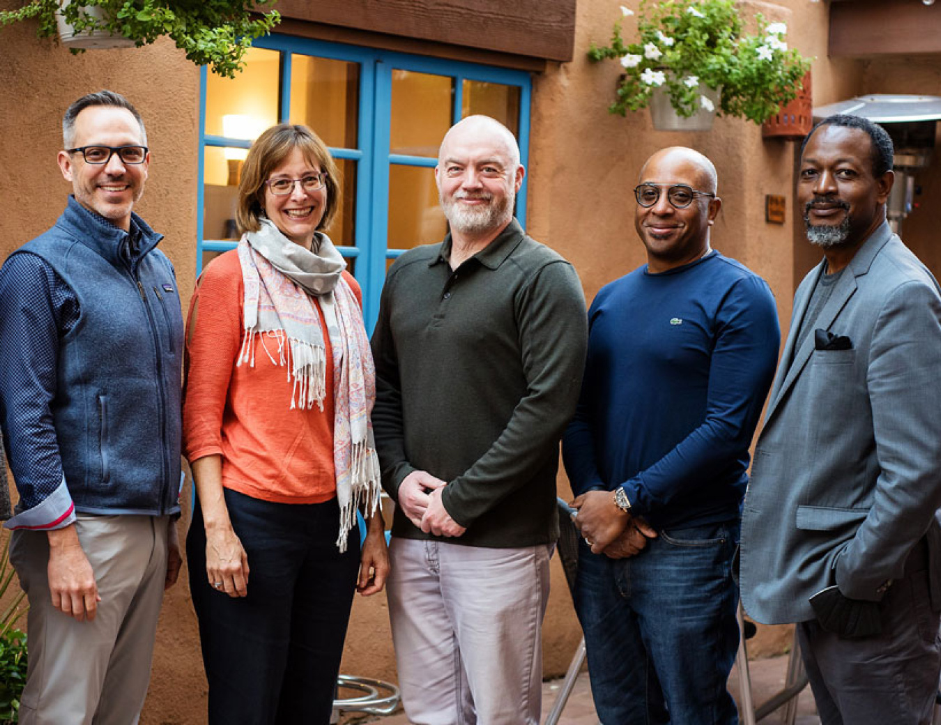Five adults standing in a row in front of a stucco building with hanging flower pots.