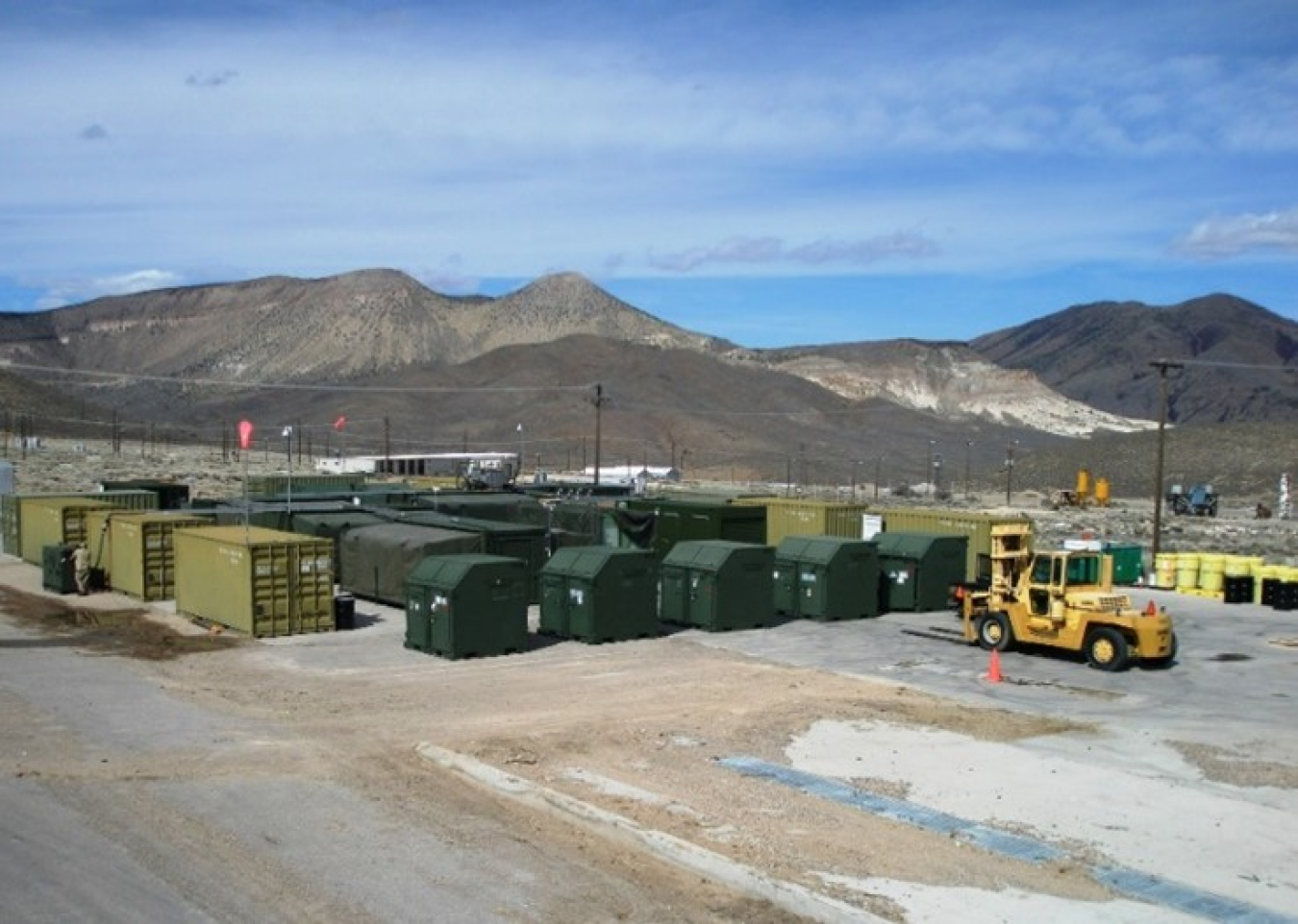 Shipping containers are lined up in a desert environment. A forklift-type loader approaches them from the right. In the background are mountains.