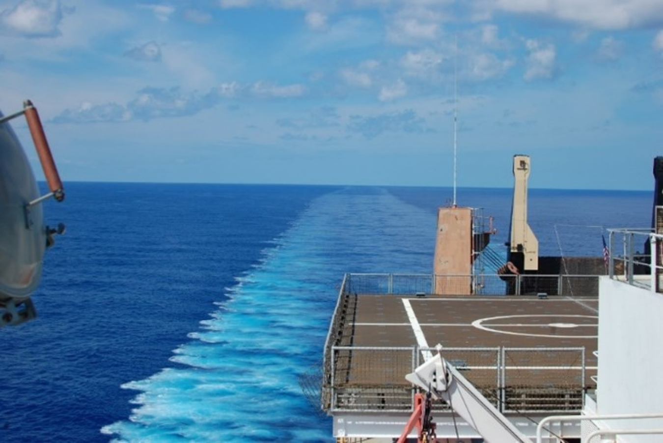 Looking off the aft of a ship while it's underway at sea. Parts of the ship are visible on the right. The wake of the ship continues into the horizon.
