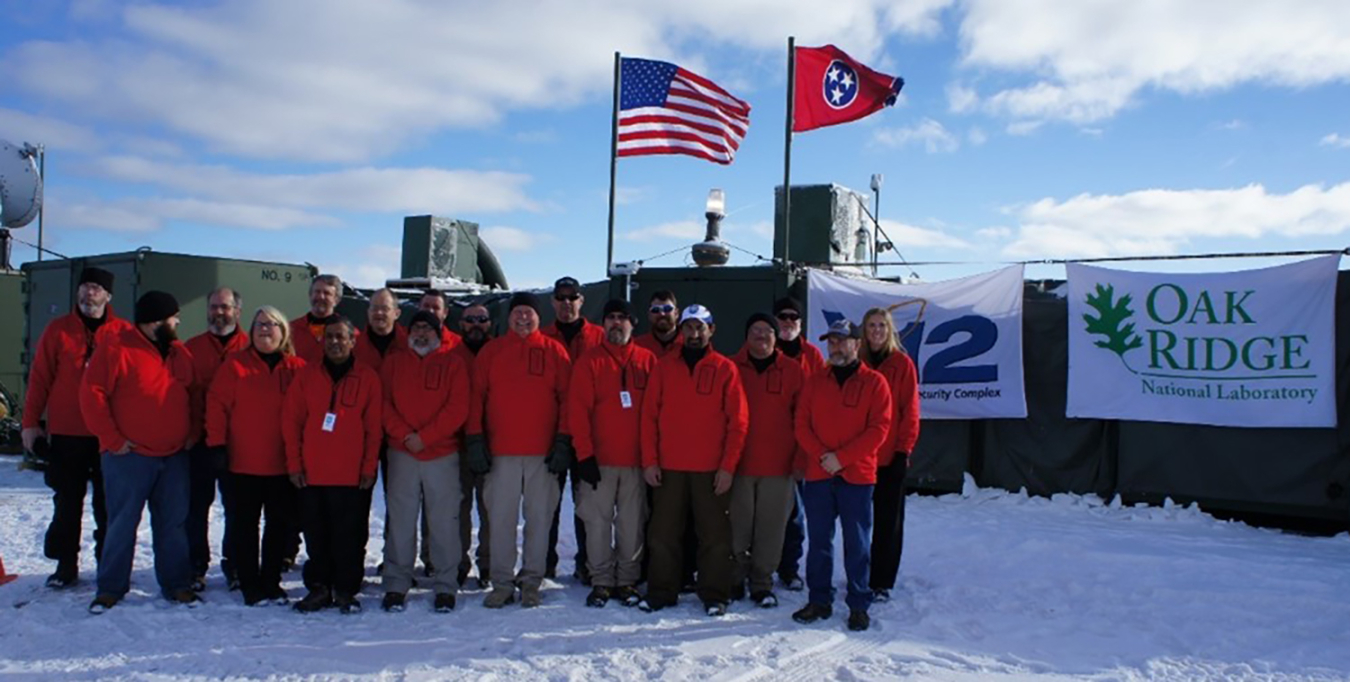 A group of people stand outside in the snow wearing matching red jackets. A U.S. and Tennessee flag fly in the background. On a wall behind them are banners with the Y-12 National Security Complex and Oak Ridge National Laboratory logos on them.