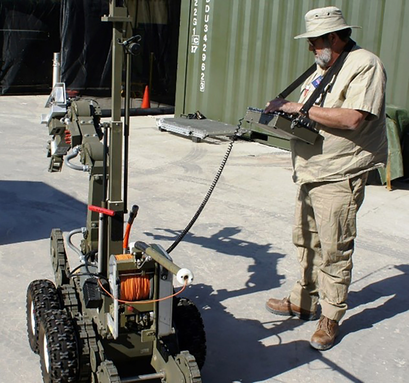 A man in short sleeves and a hat operates a robot attached by cable to a controller he is holding.