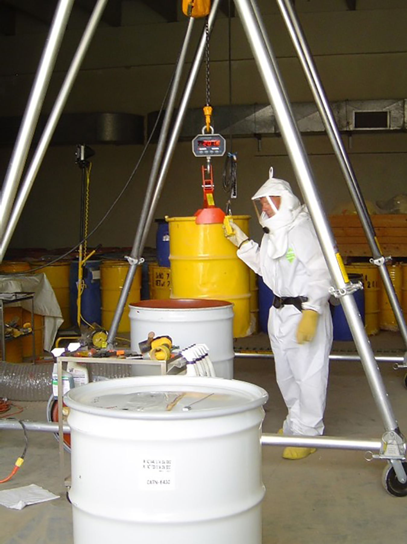A person in a "bunny suit" and gloves weighs a yellow barrel. Two white barrels sit nearby.