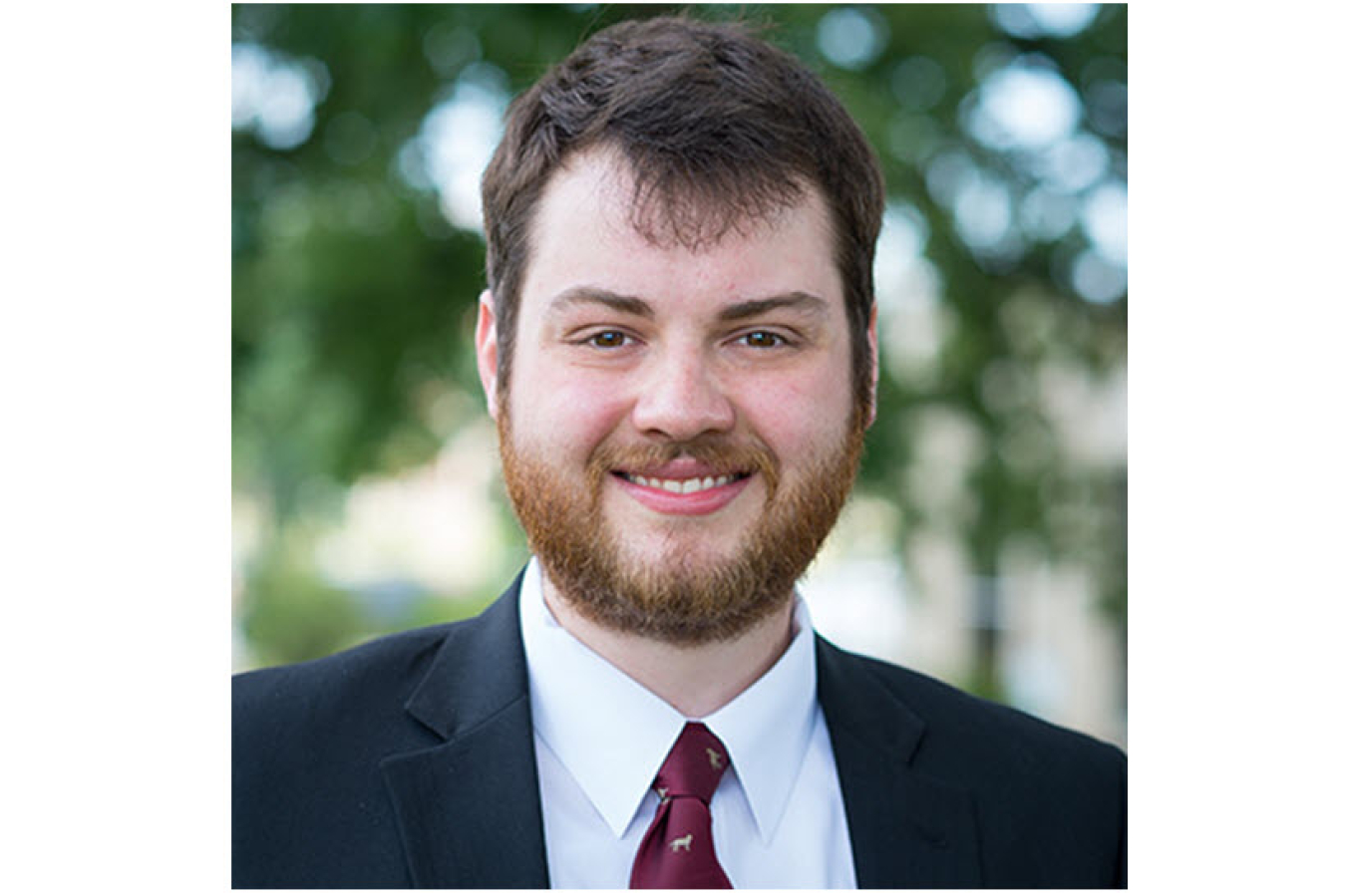 Headshot of a young man with trees in the background. He is wearing a black suit with a white shirt and red tie and smiling at the camera.
