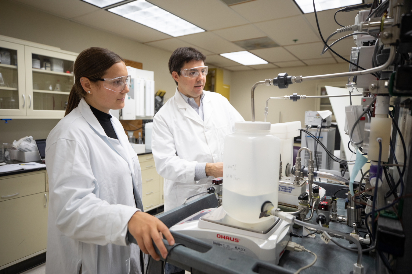 Two scientists wearing lab coats and goggles work together at a laboratory bench