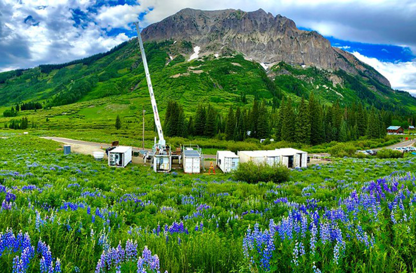 Photo of a mountain with large white containers in front of it and blue flowers in the foreground.