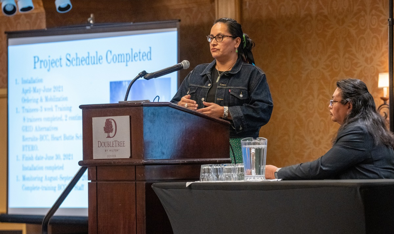 Melissa Little Plume Weatherwax speaks at the 2022 Annual Program Review (standing at podium with presentation in background) on the clean energy project at Blackfeet Community College. 