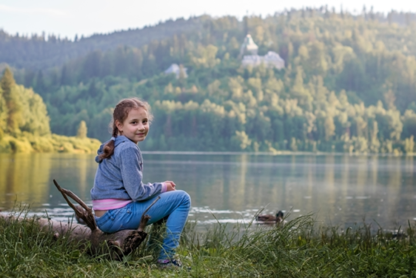 A young person sits on the side of a lake looking at the camera smiling.