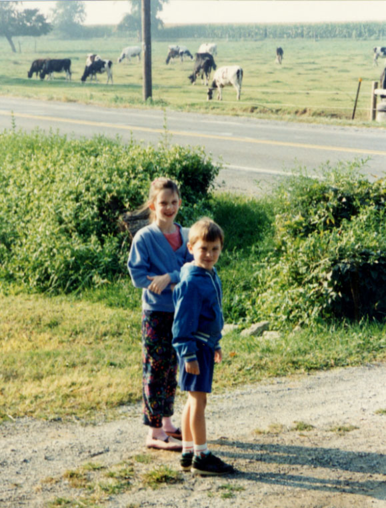 Jake Herb and his sister as young kids, standing on a dirt road near a field with cows grazing.