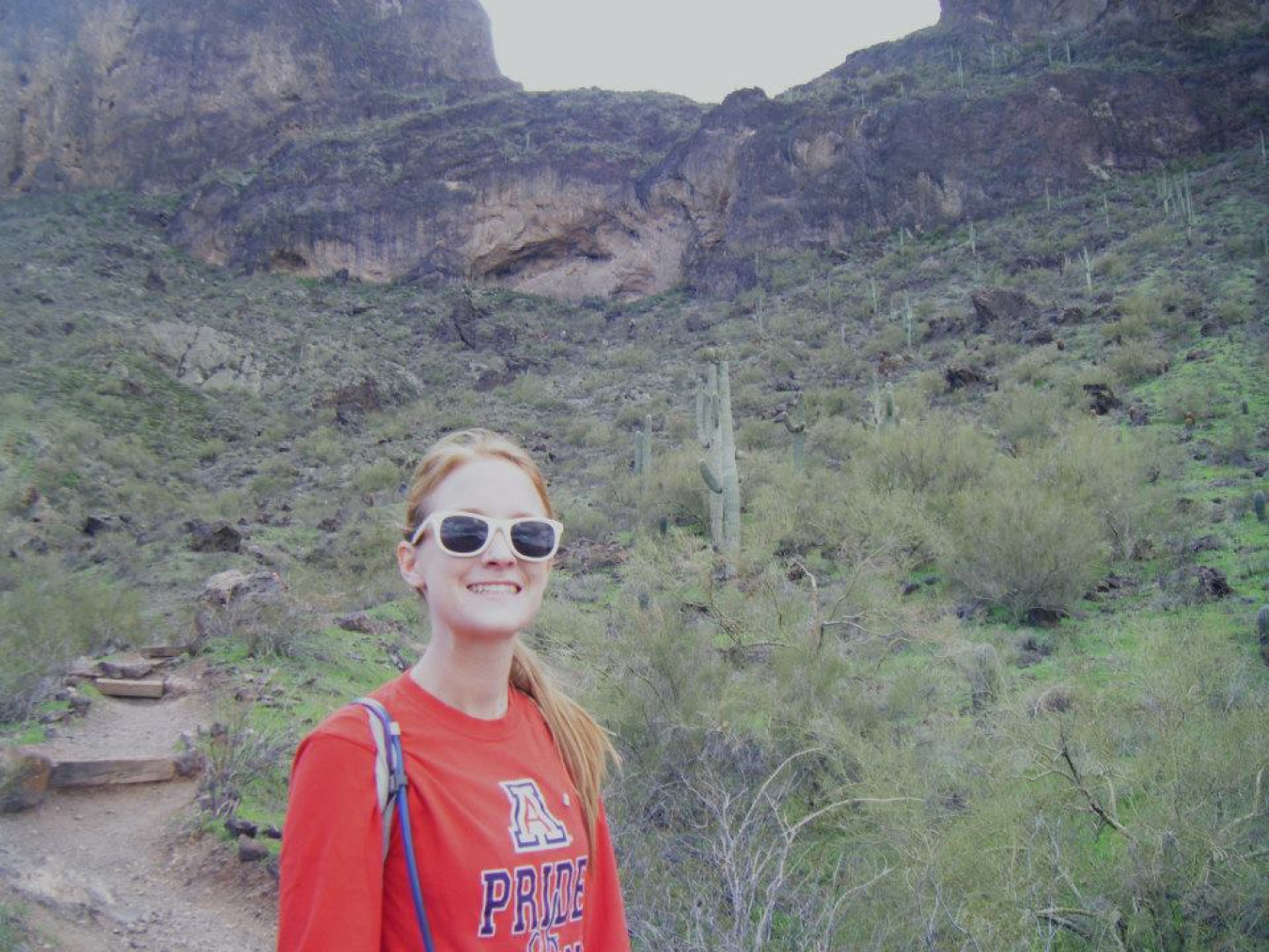 Sarah Moore standing on a trail in Arizona with cliffs, cacti, and scrub brush behind her