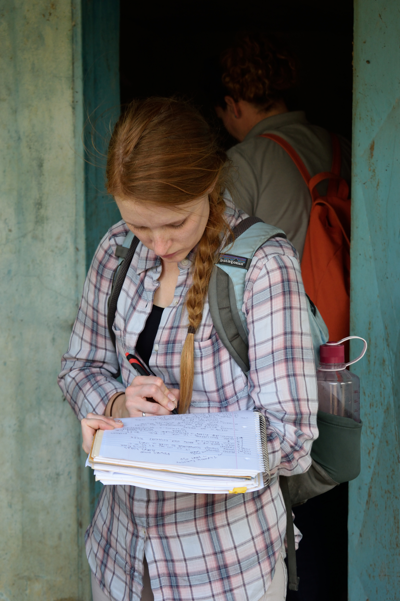 Sarah Moore, wearing a backpack and a flannel shirt, writes in a notebook that rests on her forearm.