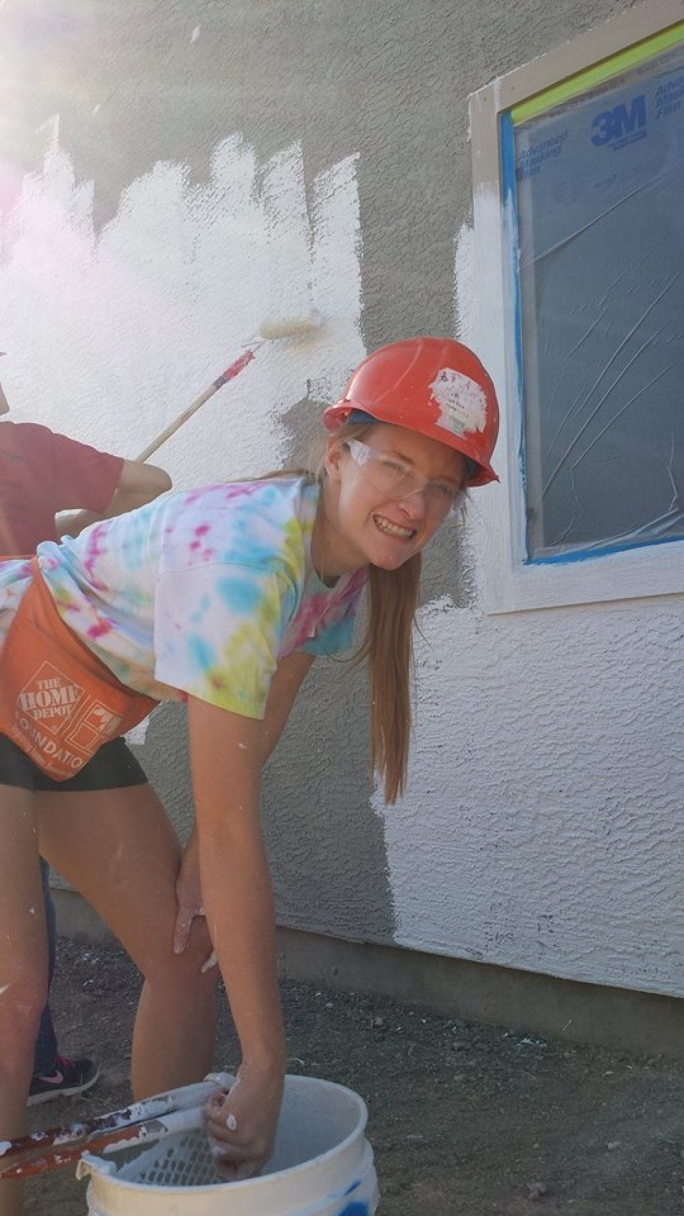 Moore, wearing a hard hat, goggles, and tie dye T-shirt, dips a paintbrush into a bucket of paint