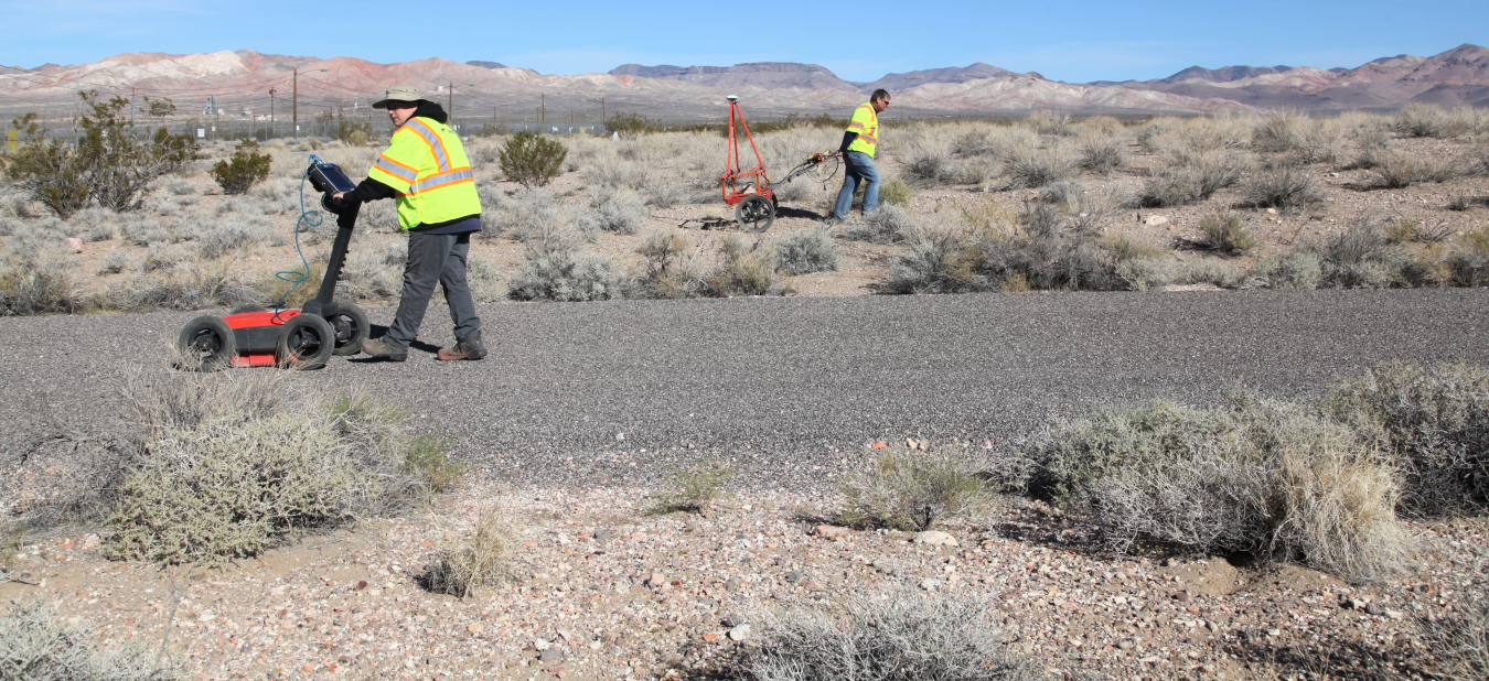 Geophysicist Beth Williams pushes a ground penetrating radar while geophysicist Jeff Warren uses a metal detector near the Engine Maintenance, Assembly, and Disassembly Facility at the Nevada National Security Site.