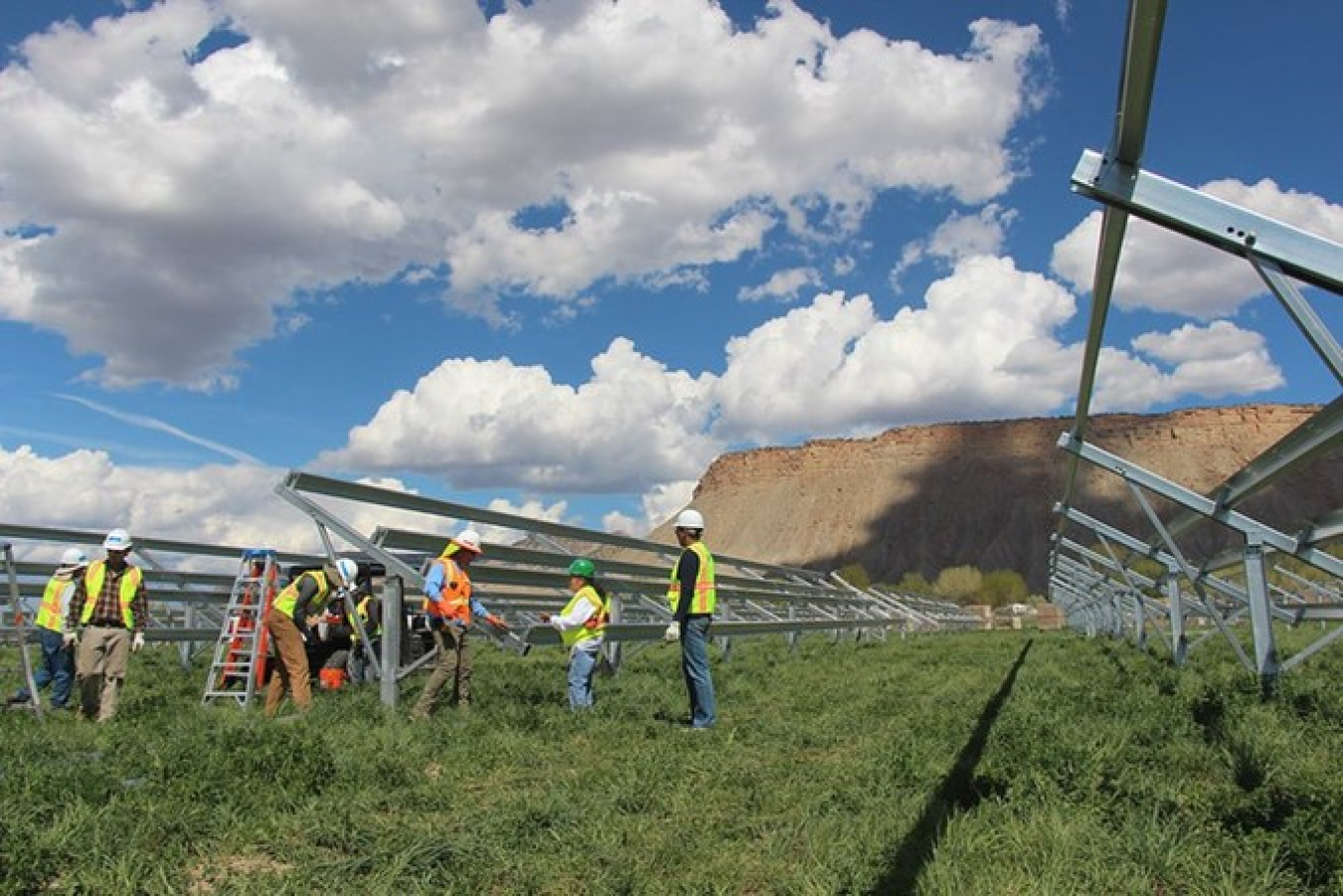 Construction workers work on installing a solar rack with a desert mesa in the background and a cloudy blue sky above.