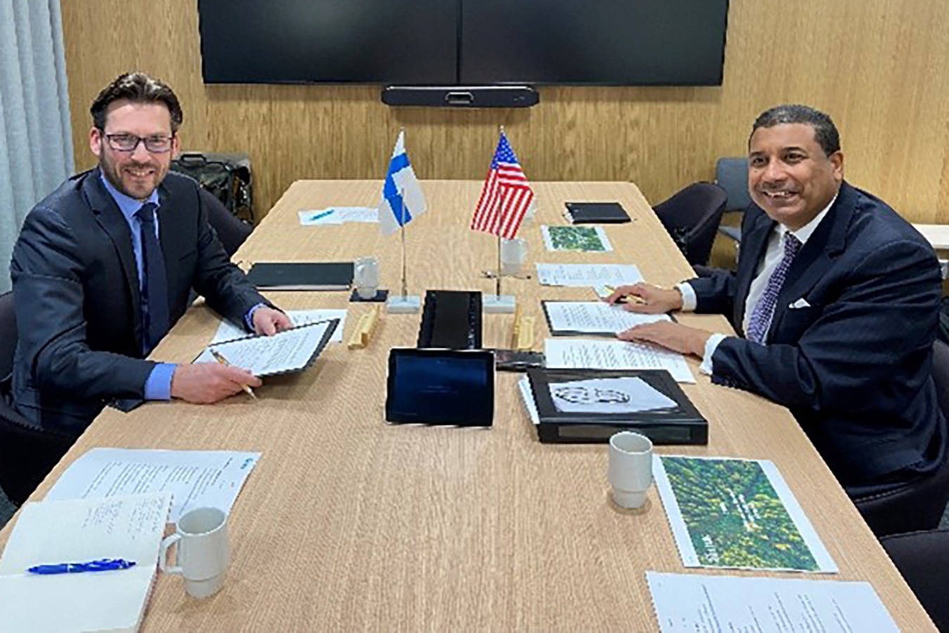 Two men sit at a long table smiling for the camera. U.S. and Finnish flags are in front of them.