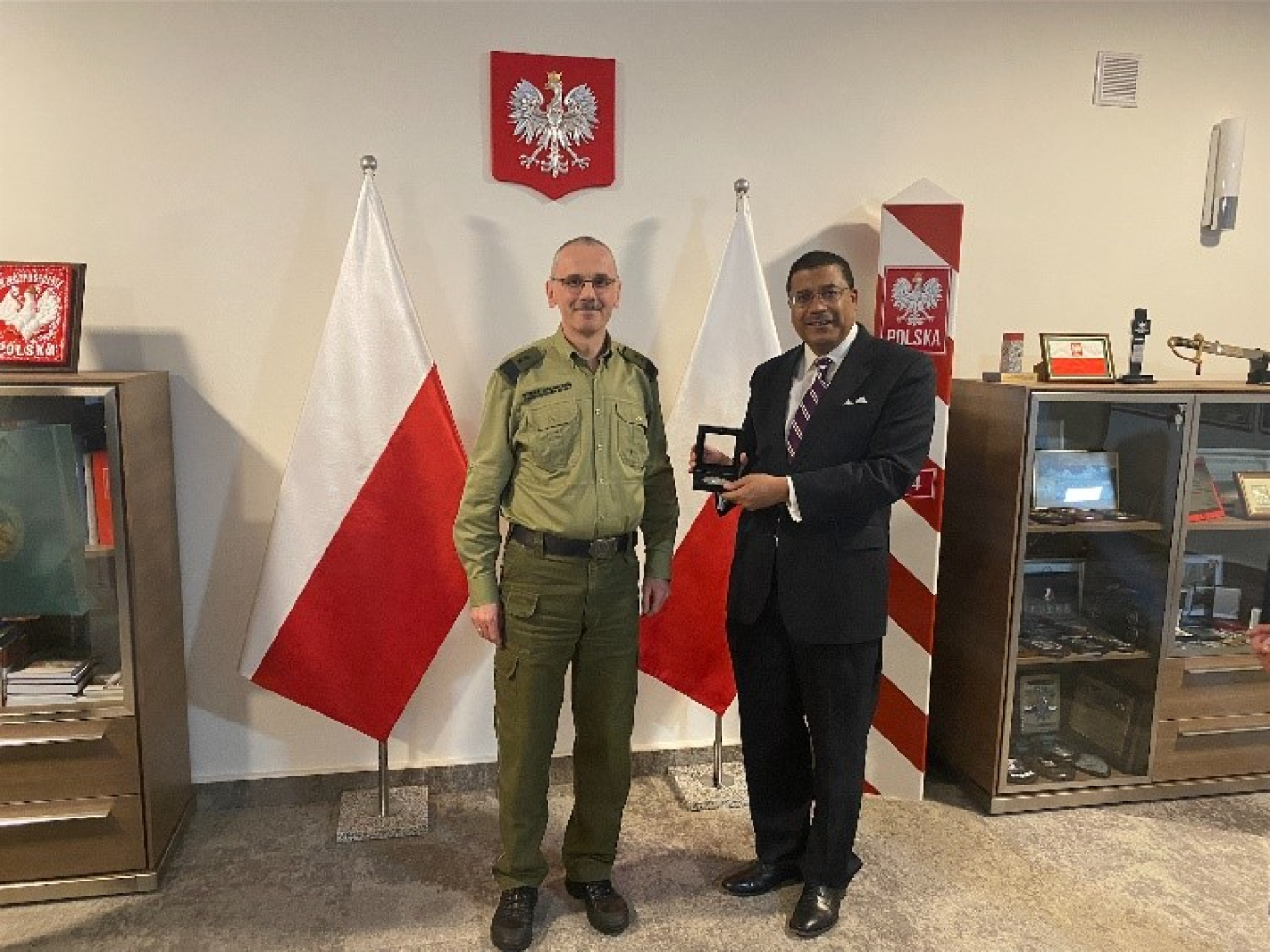 Rose and Praga stand in front of Polish flags. A coat of arms is hanging on the wall behind them.