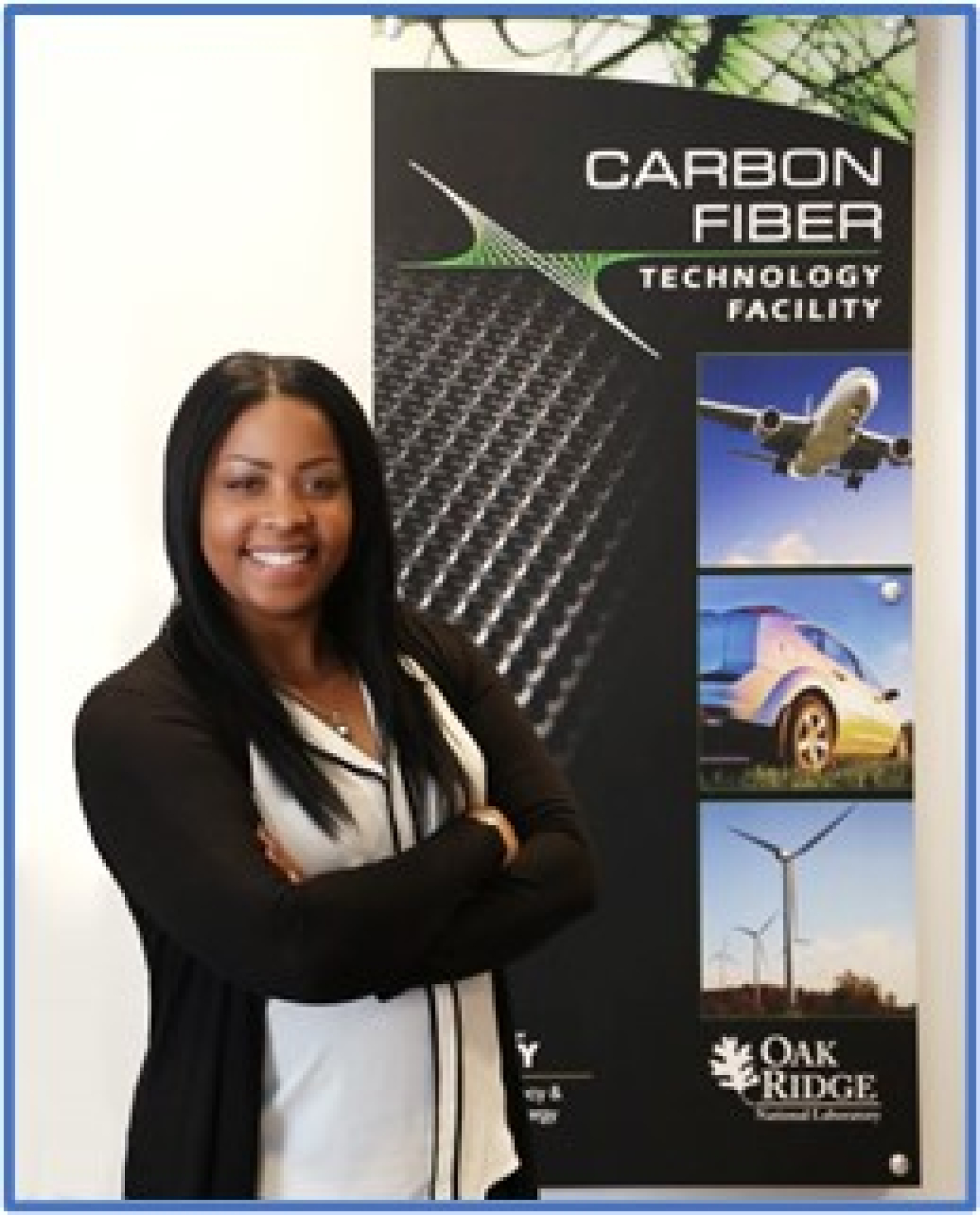 (5)	A vertical photograph of a woman standing in front of a sign that is roughly the same size as her. The woman has dark skin and long, straight dark hair. Her arms are crossed and she is smiling, wearing a white shirt and a black sweater. The sign behind her says “Carbon Fiber Technology Facility” at the top and has 3 large photos of a plane flying, a car’s rear headlight, and a wind turbine in a large field. At the bottom of the sign is a large logo for Oak Ridge National Laboratory. 