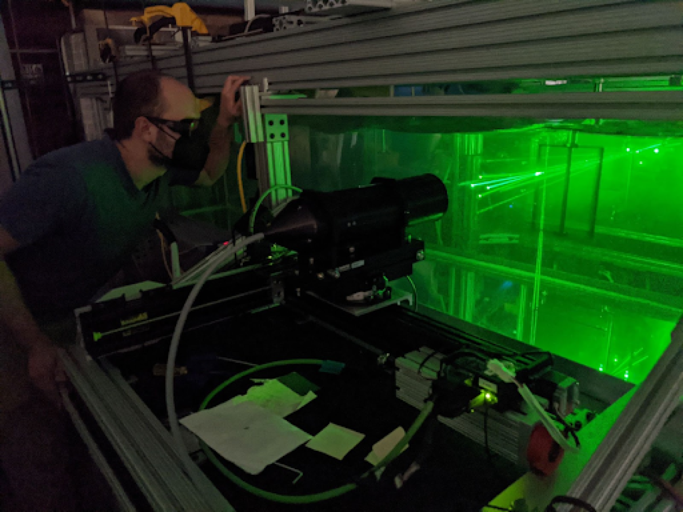 Person with eye protection looking at green beams of light in a contained clear box.