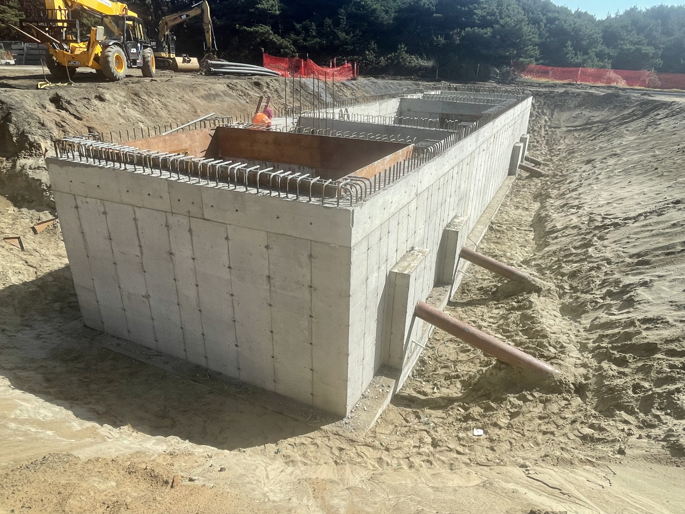 Photo of a rectangular cement structure on top of sand, with an open top showing rebar and four conduit pipes angling down into the sand at the side.