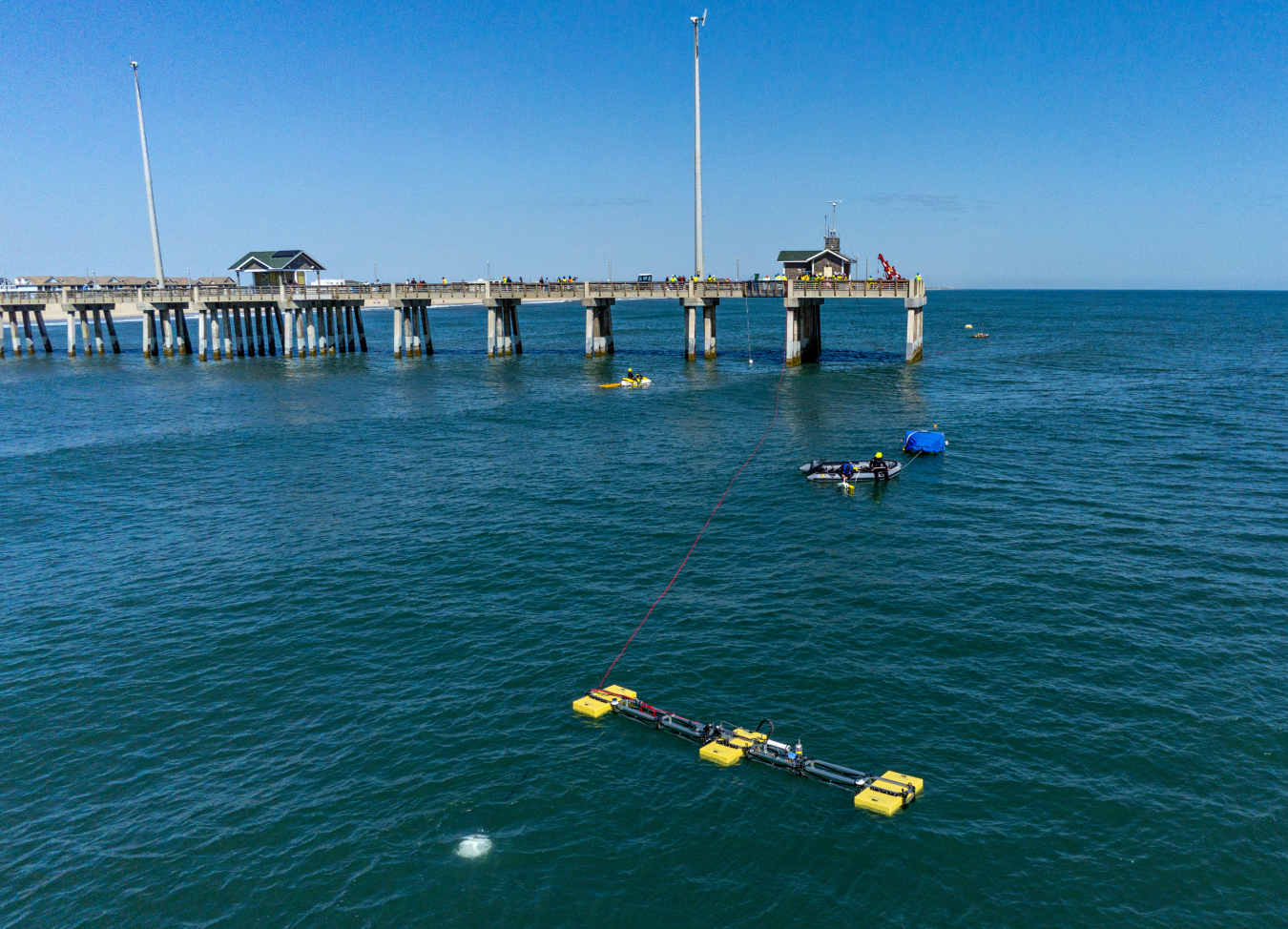 A photo of wave-powered desalination devices, boats, and people in the ocean near a pier.