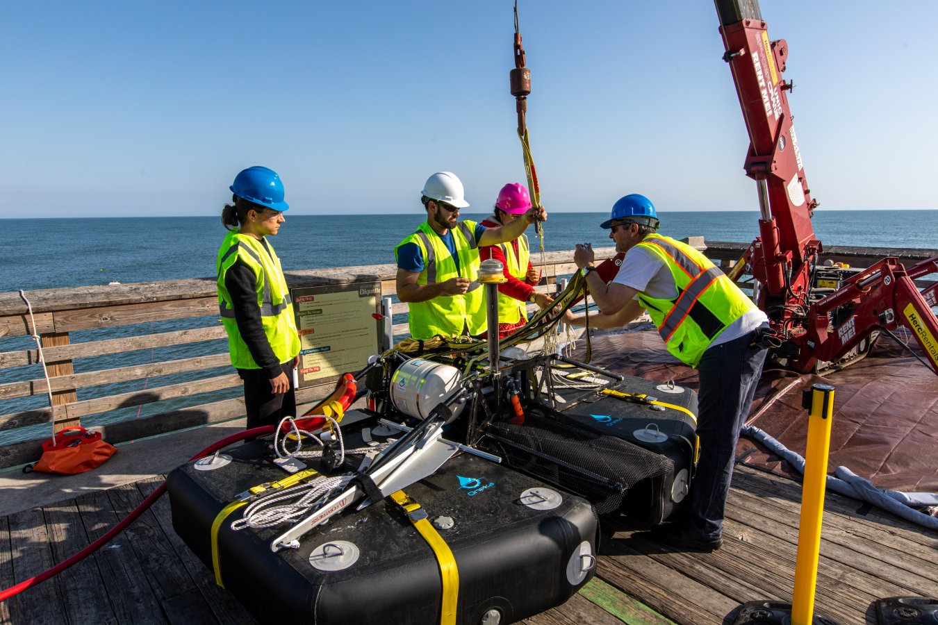 A group of people wearing yellow safety vests and hard hats secure a wave-powered desalination prototype to a crane.