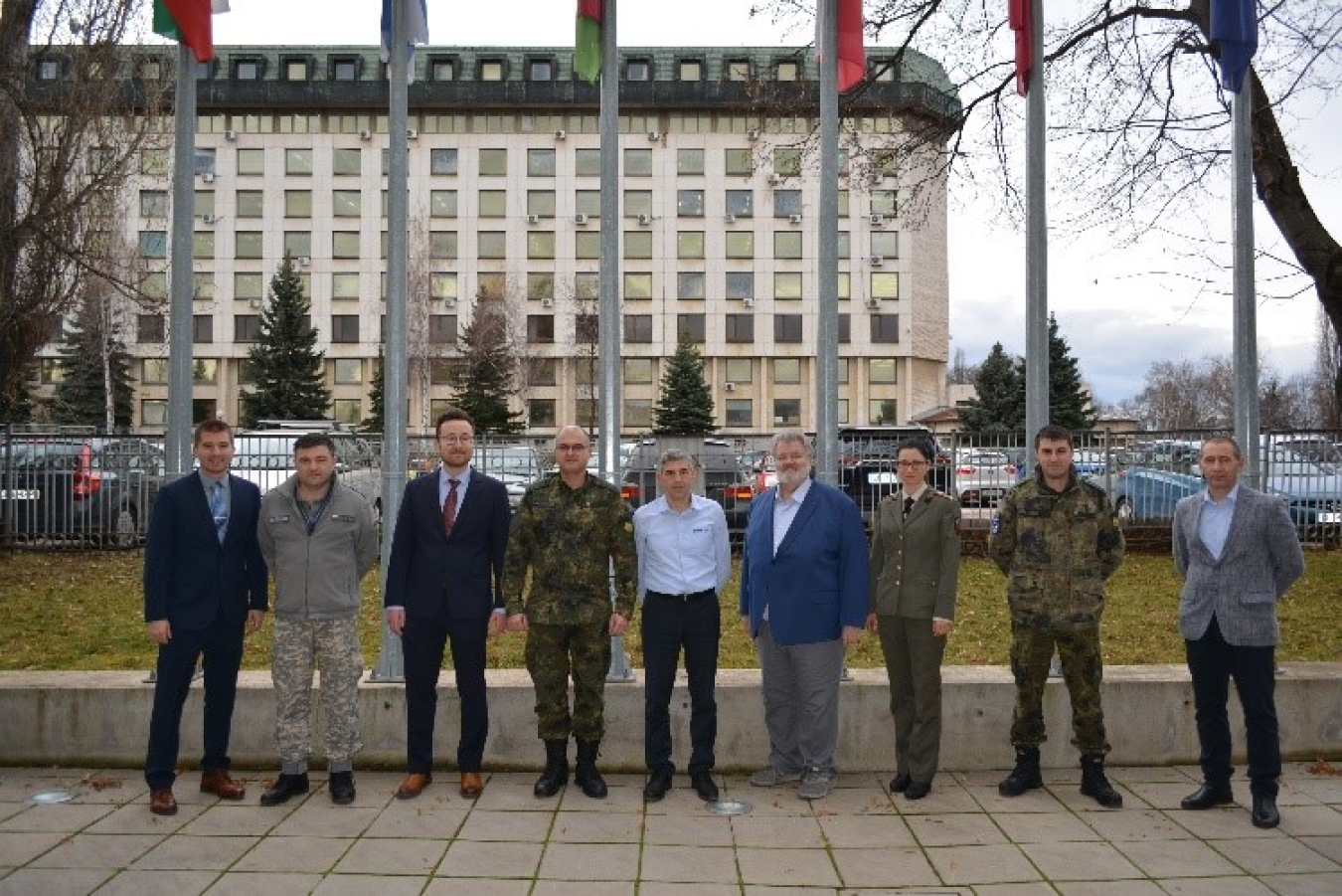 A photo of eight men and a woman -- some in uniform -- standing in front of a row of flagpoles.