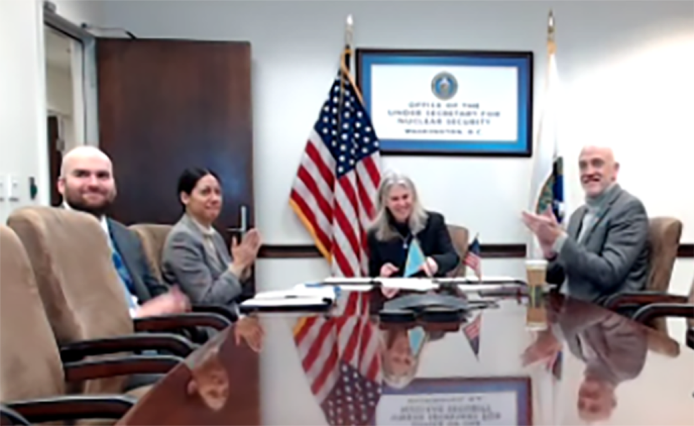 Administrator Hruby signs a document while smiling at a virtual meeting. Three NNSA staffers sit next to her, clapping.