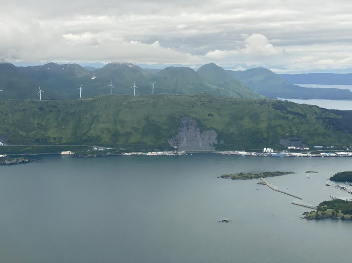 A body of water with a large hill with 5 wind turbines on top.