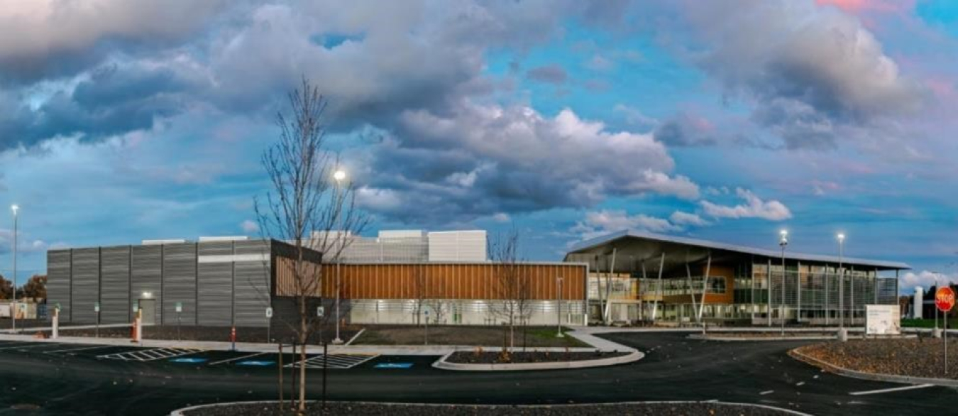 Photo of a short, long modern building with gray and brown tiling and a parking lot with trees in front of it.