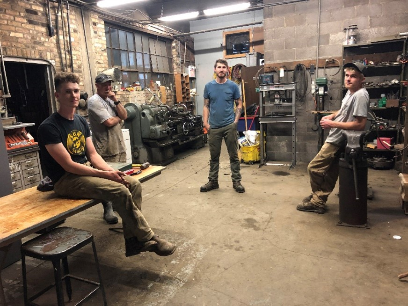Four men standing or sitting in a brick-walled room with tools and mechanical equipment scattered on shelves and a table
