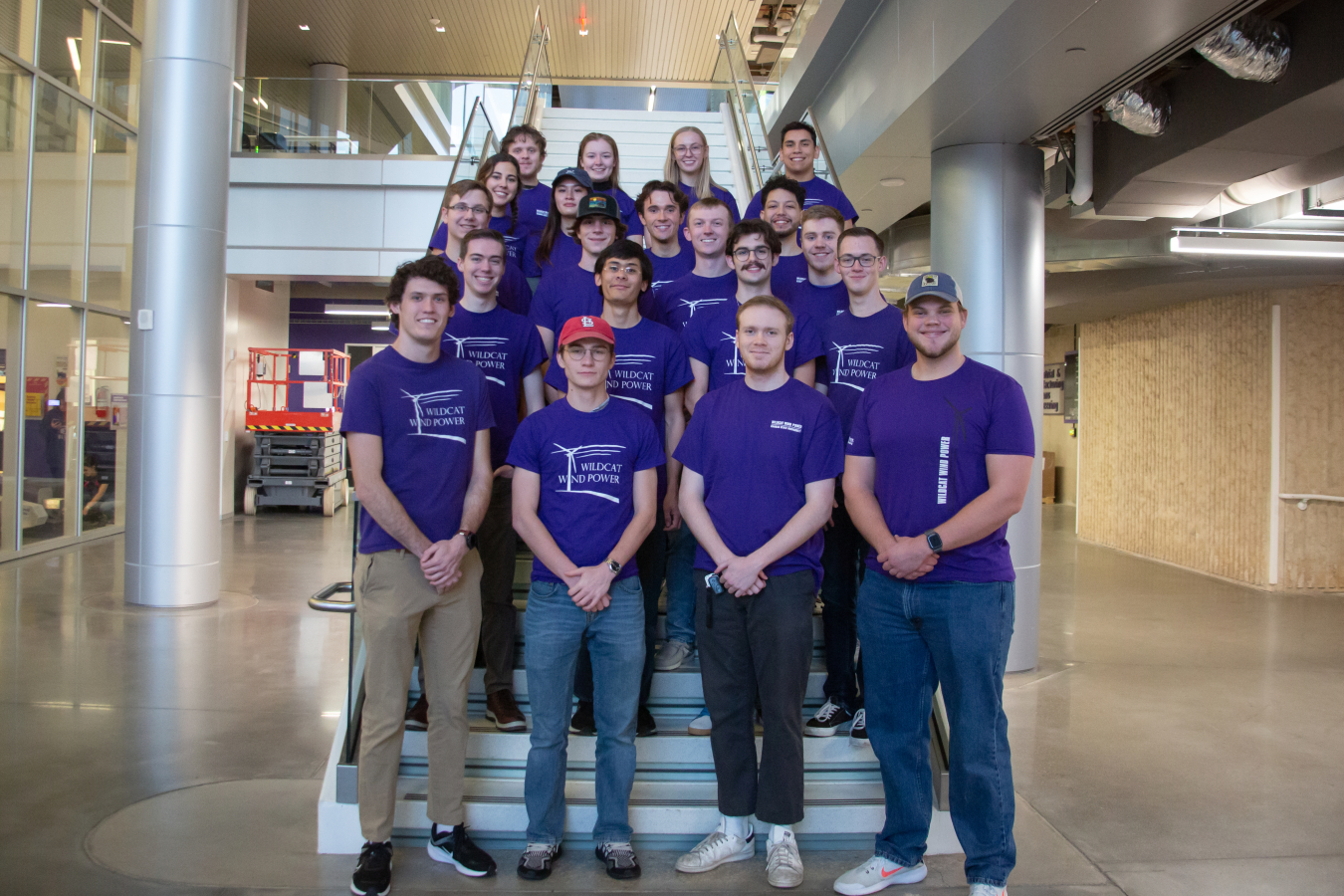 Five rows of students in matching shirts standing on stairs inside an atrium.