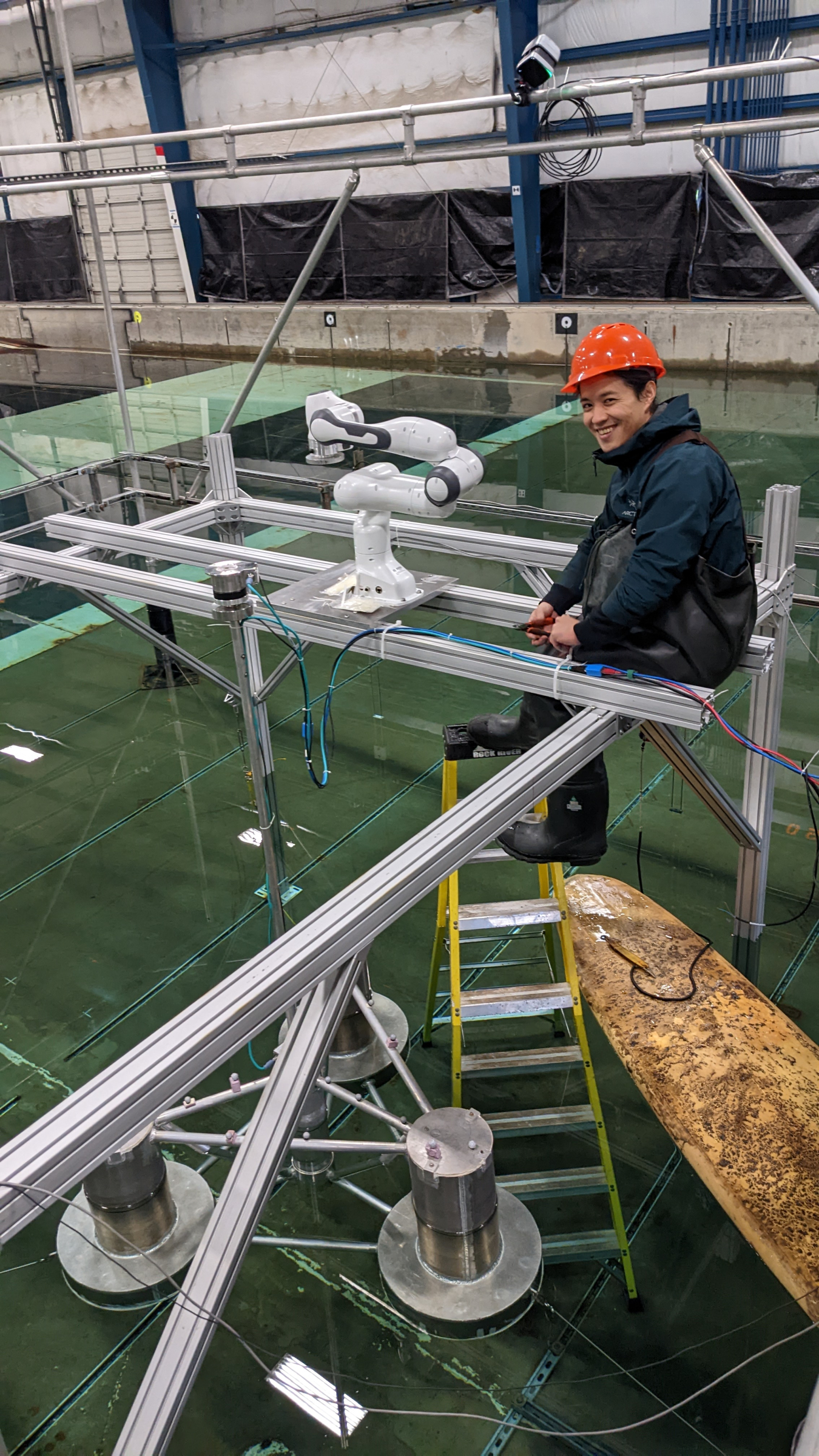 A student wearing a hard hat and waterproof attire sits on a structural beam above a wave tank. A robotic arm is attached to the beam next to him and a surfboard and offshore wind turbine tower float in the water below.