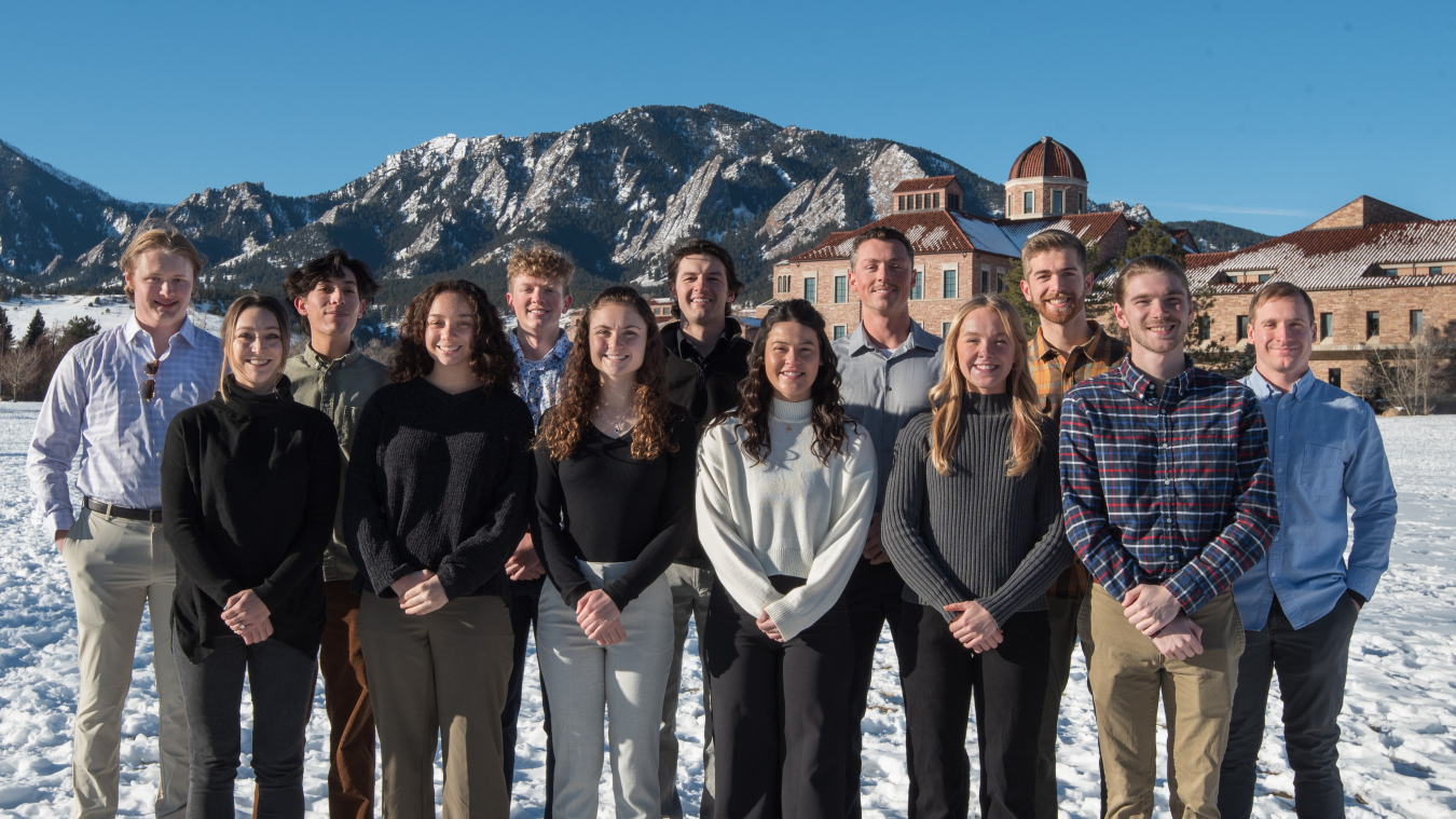 Two rows of students standing in snow in front of brick buildings and mountains.