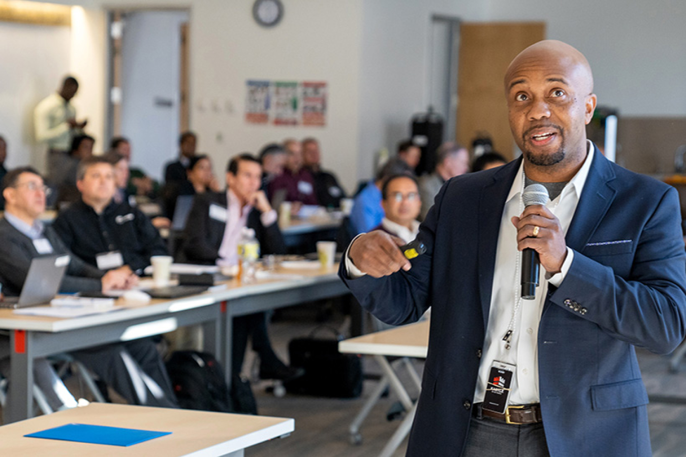 Man in a suit holding a microphone addressing a room full of people.
