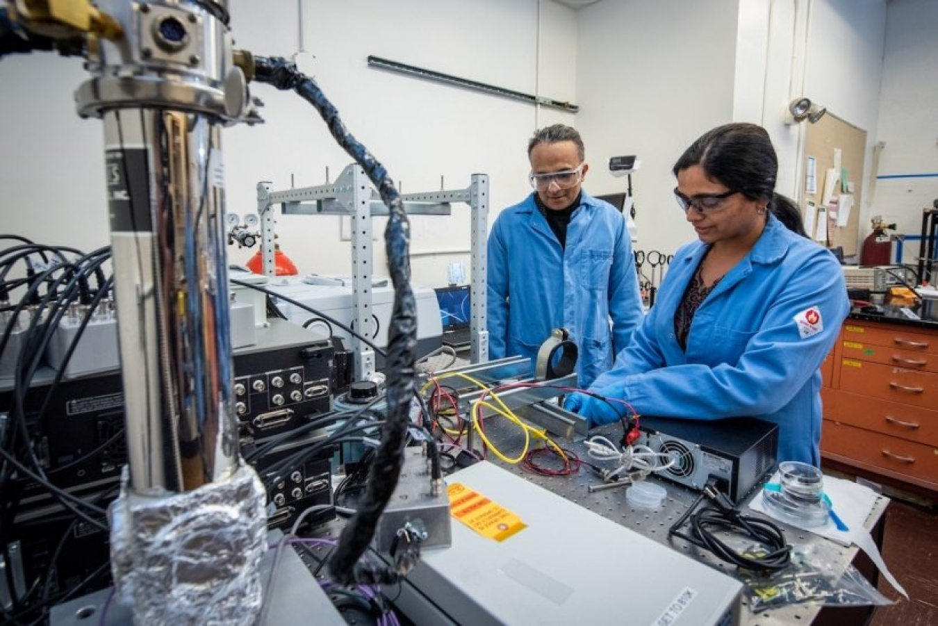 Two people wearing lab coats working in a lab; one is standing at a table working with equipment, and the other is observing.