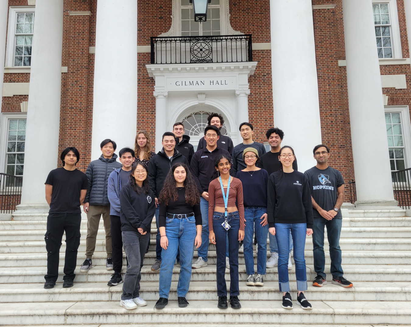 Three rows of students standing on steps in front of a large brick building labeled “Gilman Hall.”